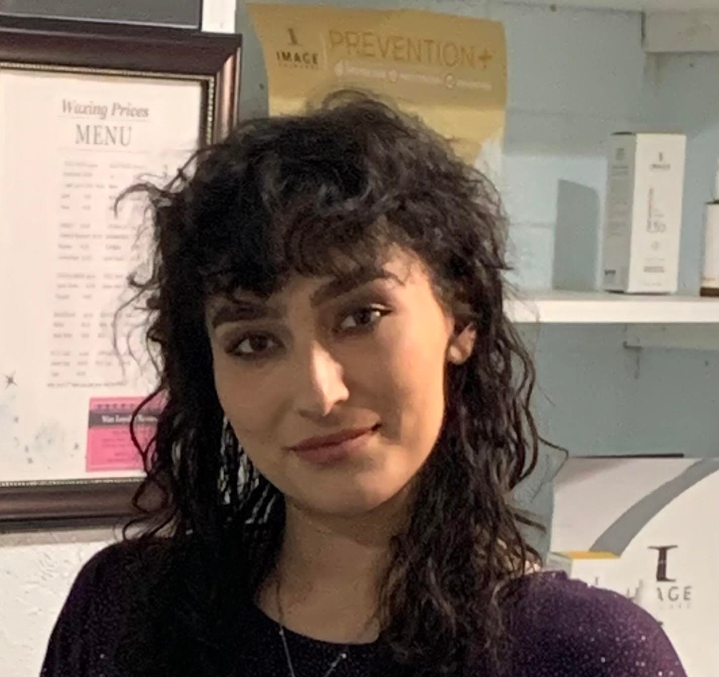 Woman with dark curly hair and bangs smiling in front of a salon menu.